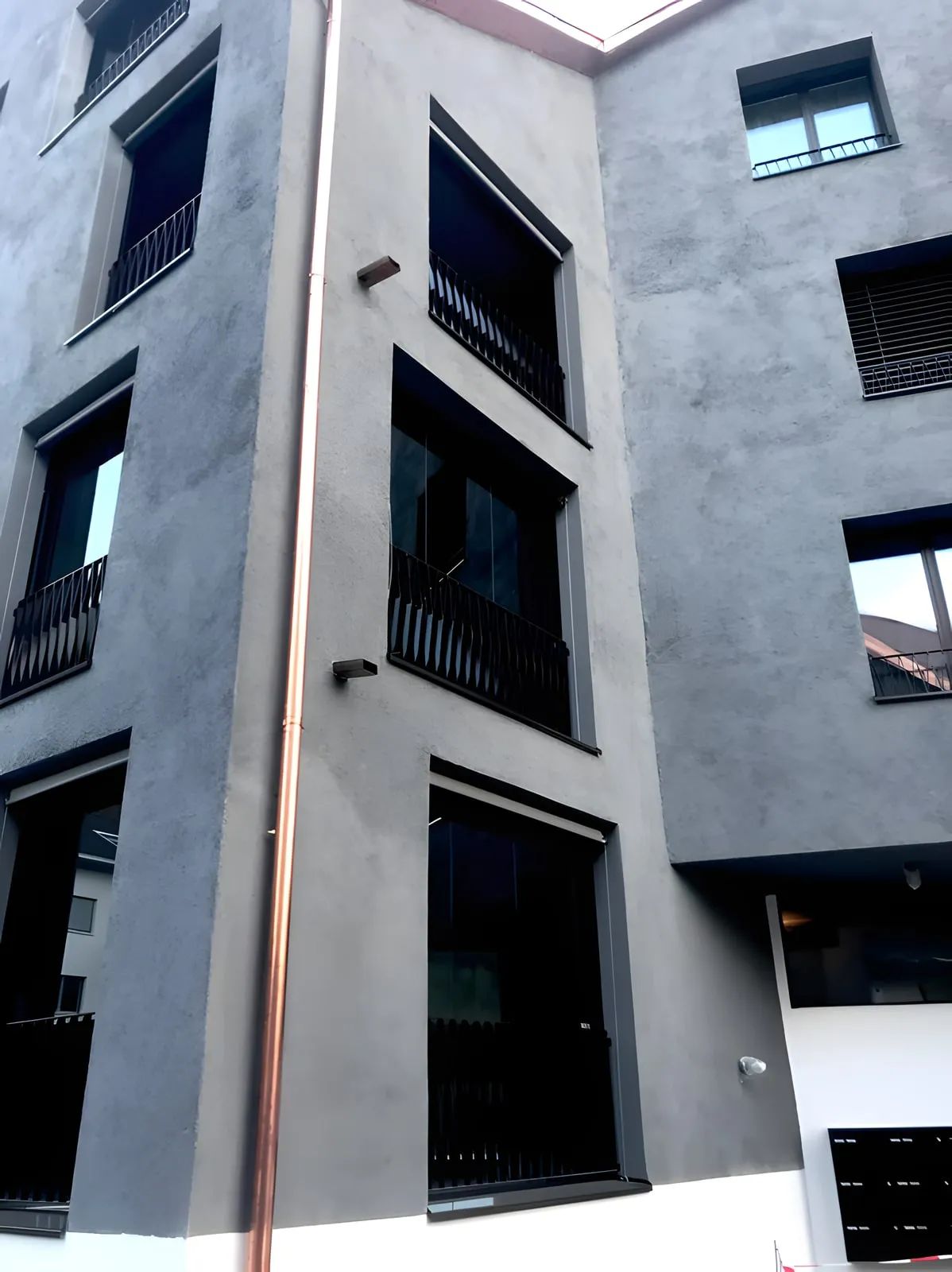 Graue Hausfassade mit dunklen Fenstern und Absturzsicherungen aus Metall
